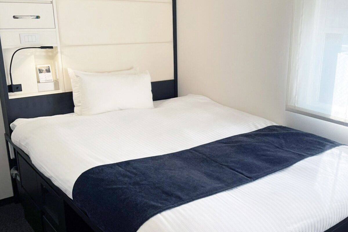 A neatly made bed with a white and dark blue bedspread, a bedside table with a lamp, and a window with blinds.