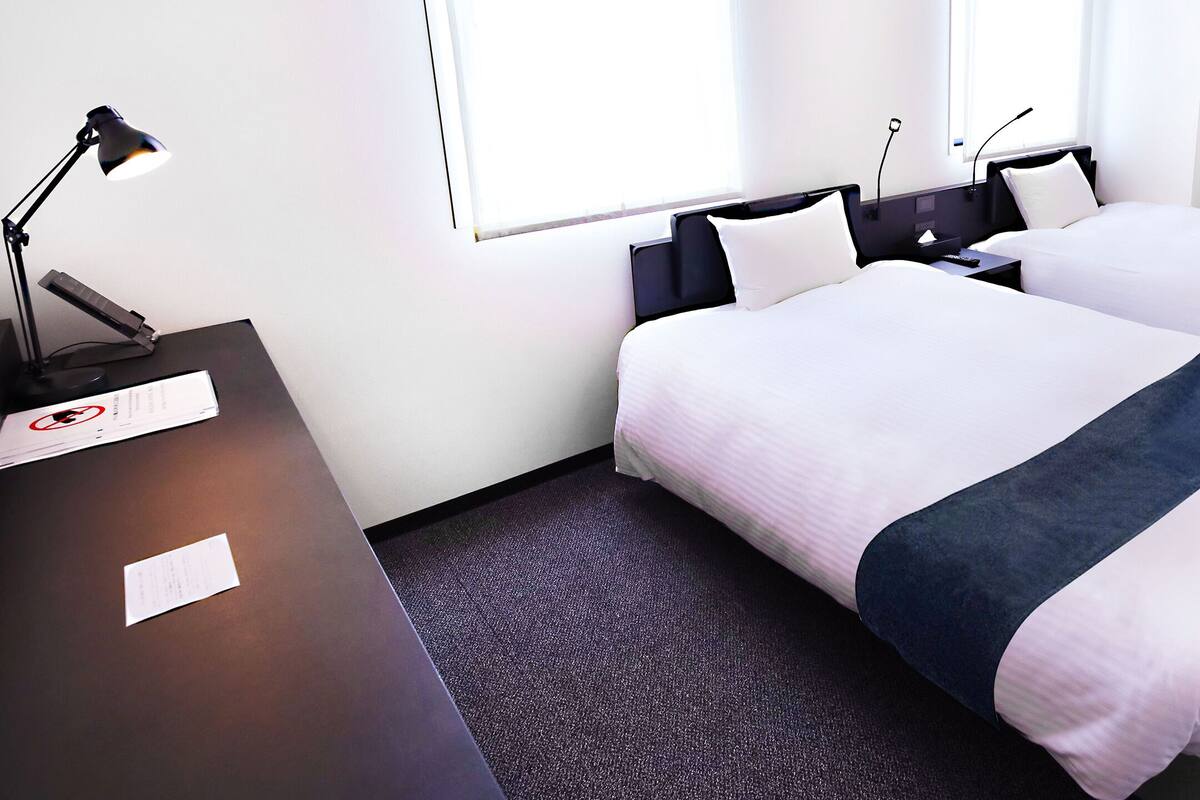 A hotel room with two beds, a desk with a lamp, and a window with blinds.