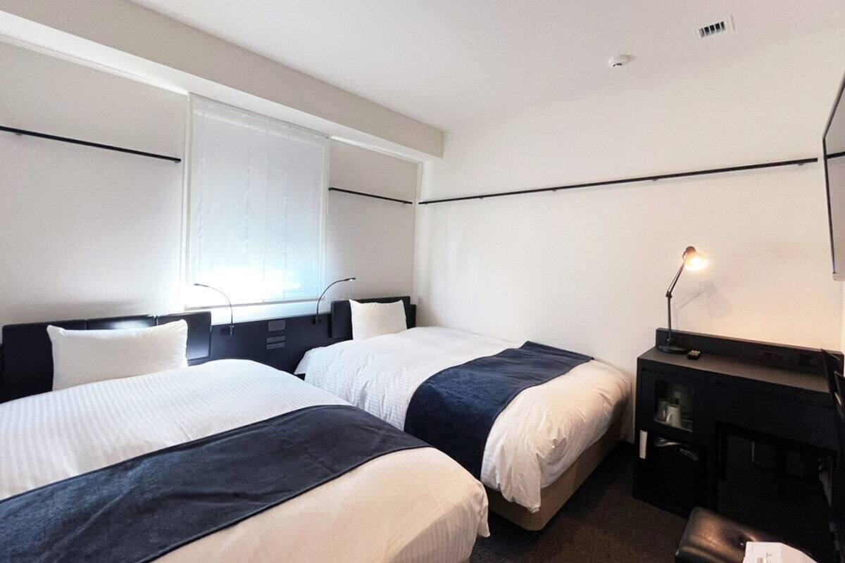 A hotel room with two beds, a desk, a chair, a lamp, and a window with blinds.