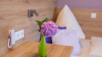 A wooden bedside table with a green vase containing pink flowers, a white pillow, and a white blanket on a bed with a wooden headboard.