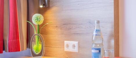 A wooden wall with a power outlet, a remote control, a glass vase with a plant, a bottle of water, and a bowl of fruit.