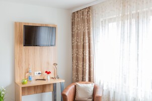 Quarto moderno com televisão na parede, secretária de madeira com um vaso de flores vermelhas e uma poltrona de couro.