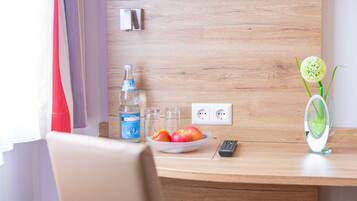 A wooden desk with a bowl of fruit, a bottle of water, glasses, a remote control, and a vase with a flower.