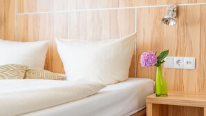 Uma cama bem arrumada com almofadas brancas e estampadas, um vaso verde com uma flor e uma mesa de cabeceira de madeira com tomada.