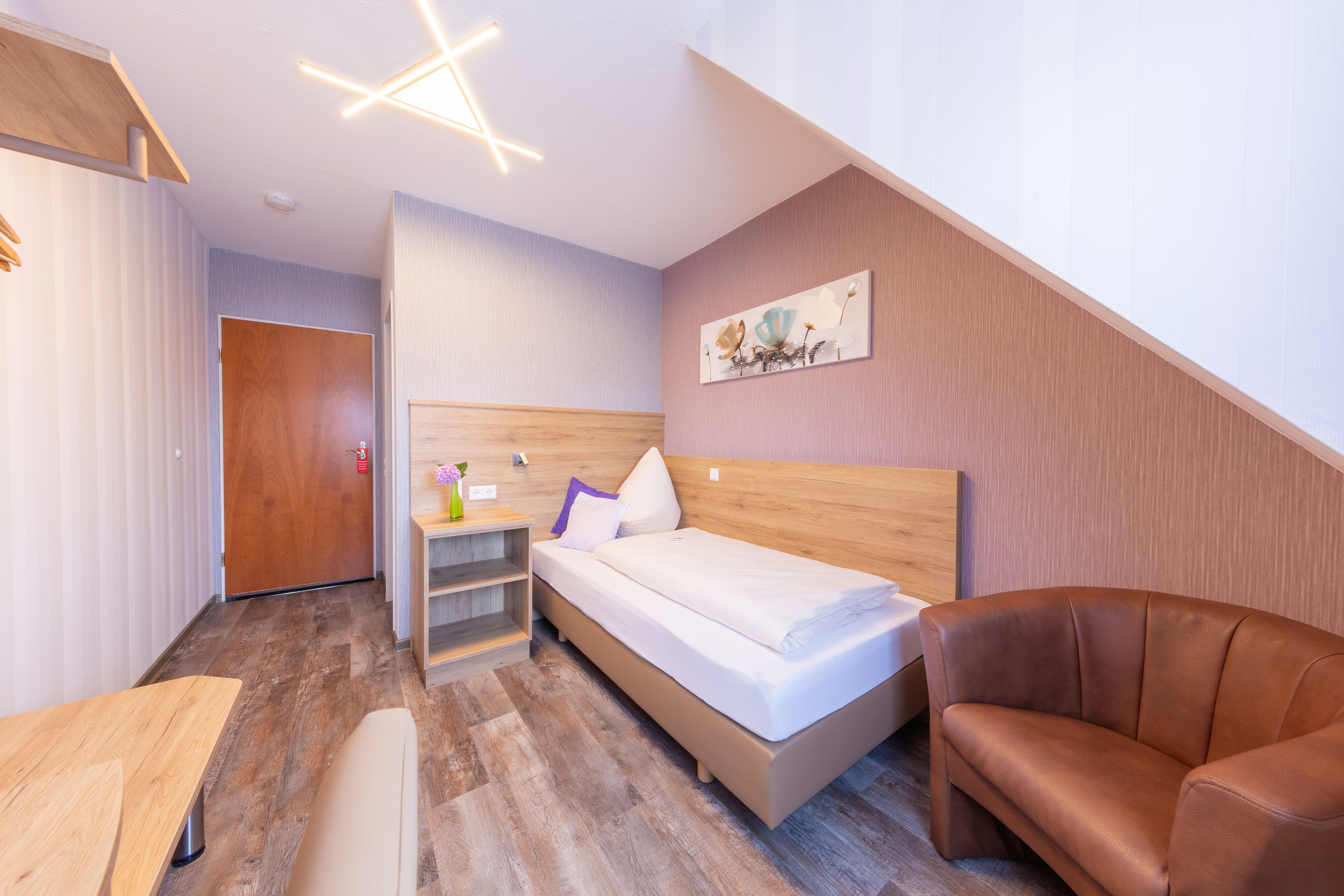 A modern hotel room with a wooden headboard, a bed with white linens, a brown leather armchair, and a small desk with a vase of flowers.