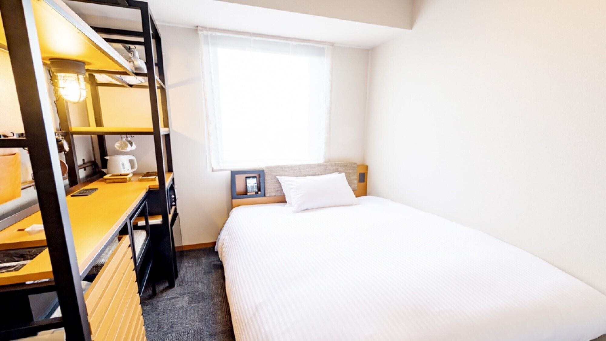 A compact hotel room with a bed, a desk with a lamp, a small table with a cup, and a window with blinds.