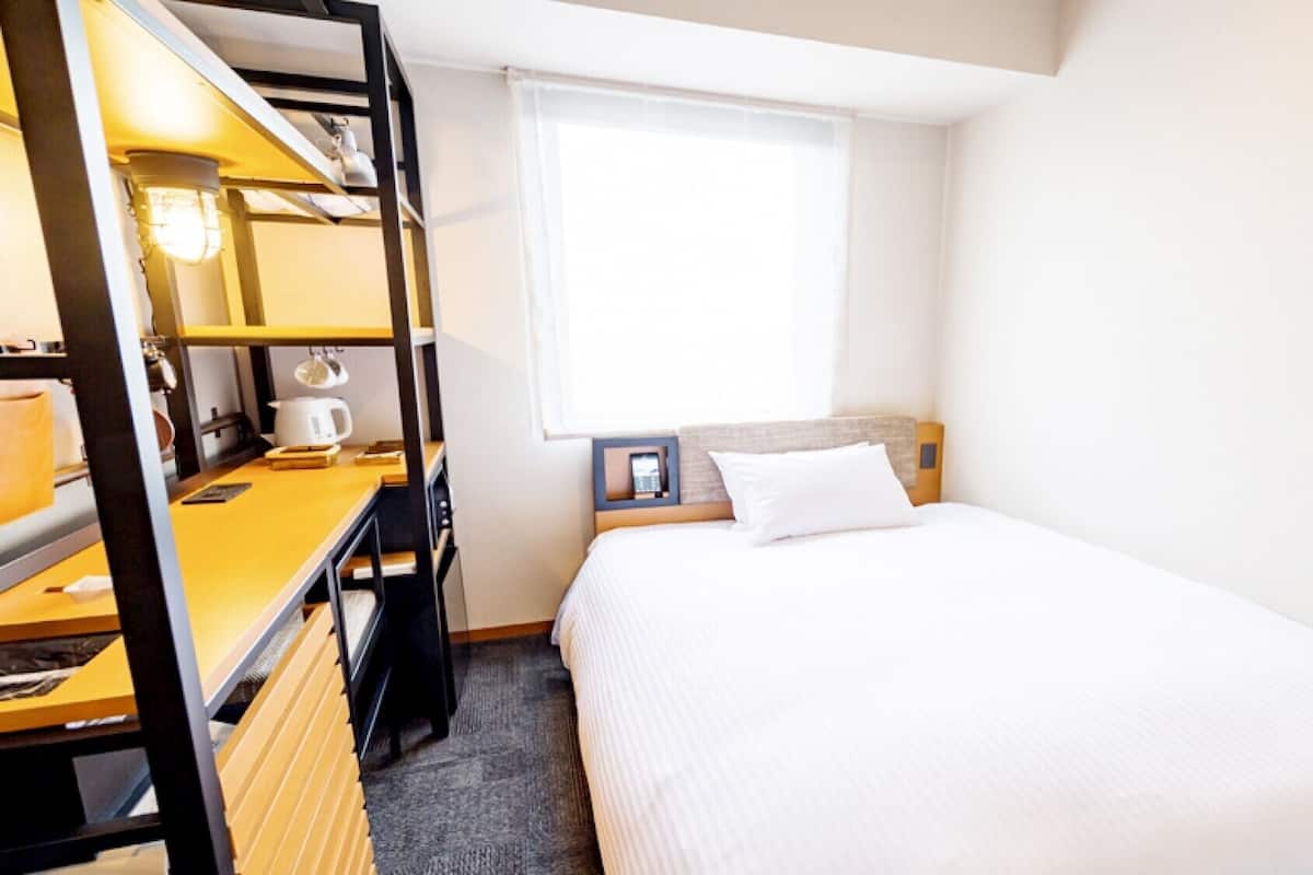 A compact hotel room with a bed, a desk with a lamp, a small table with a cup, and a window with blinds.