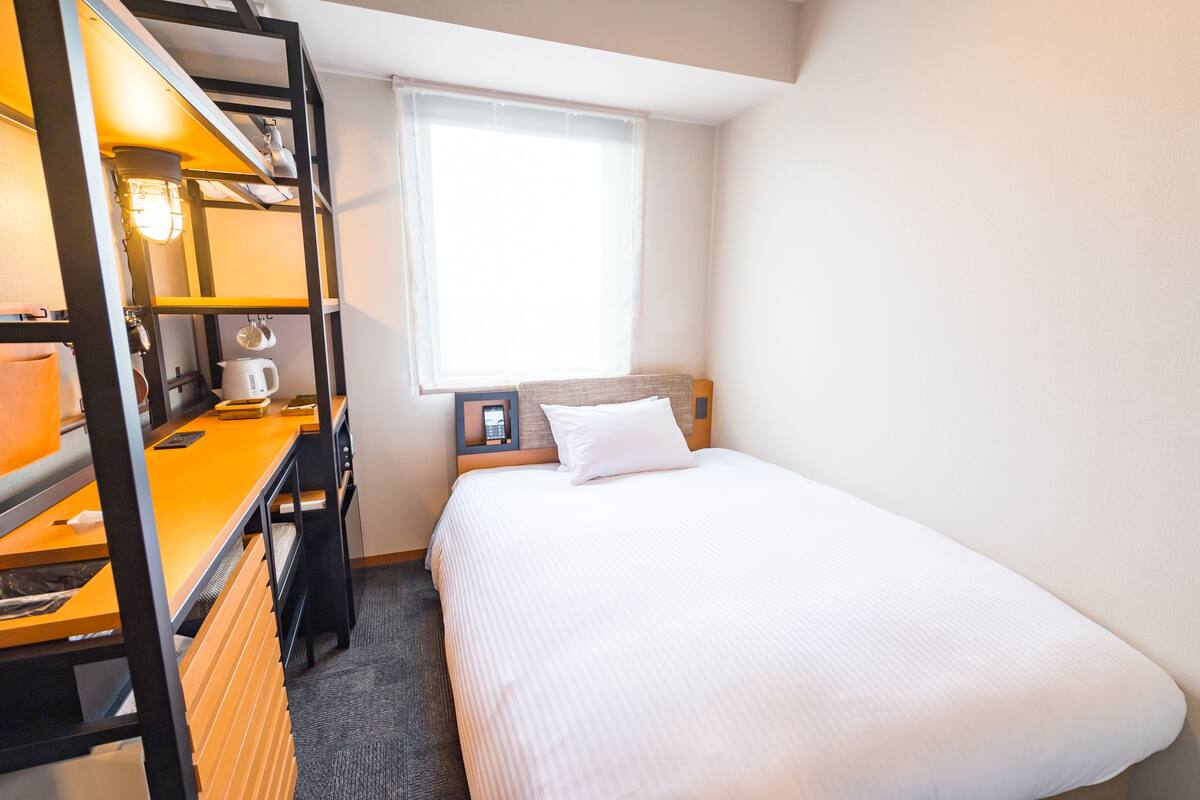 A compact hotel room with a bunk bed, a bedside table with a lamp, a small desk with a chair, and a window providing natural light.