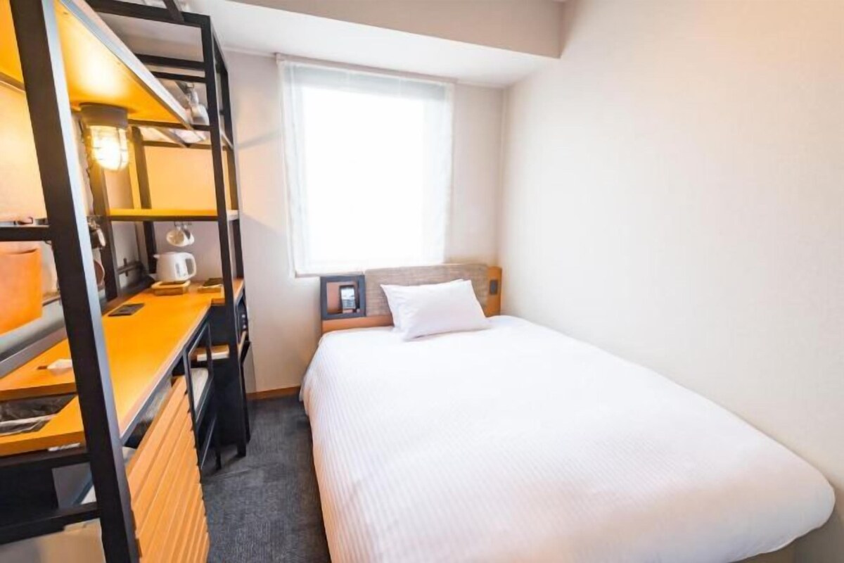 A compact hotel room with a bunk bed, a small desk with a lamp, a bedside table with a lamp, and a window providing natural light.