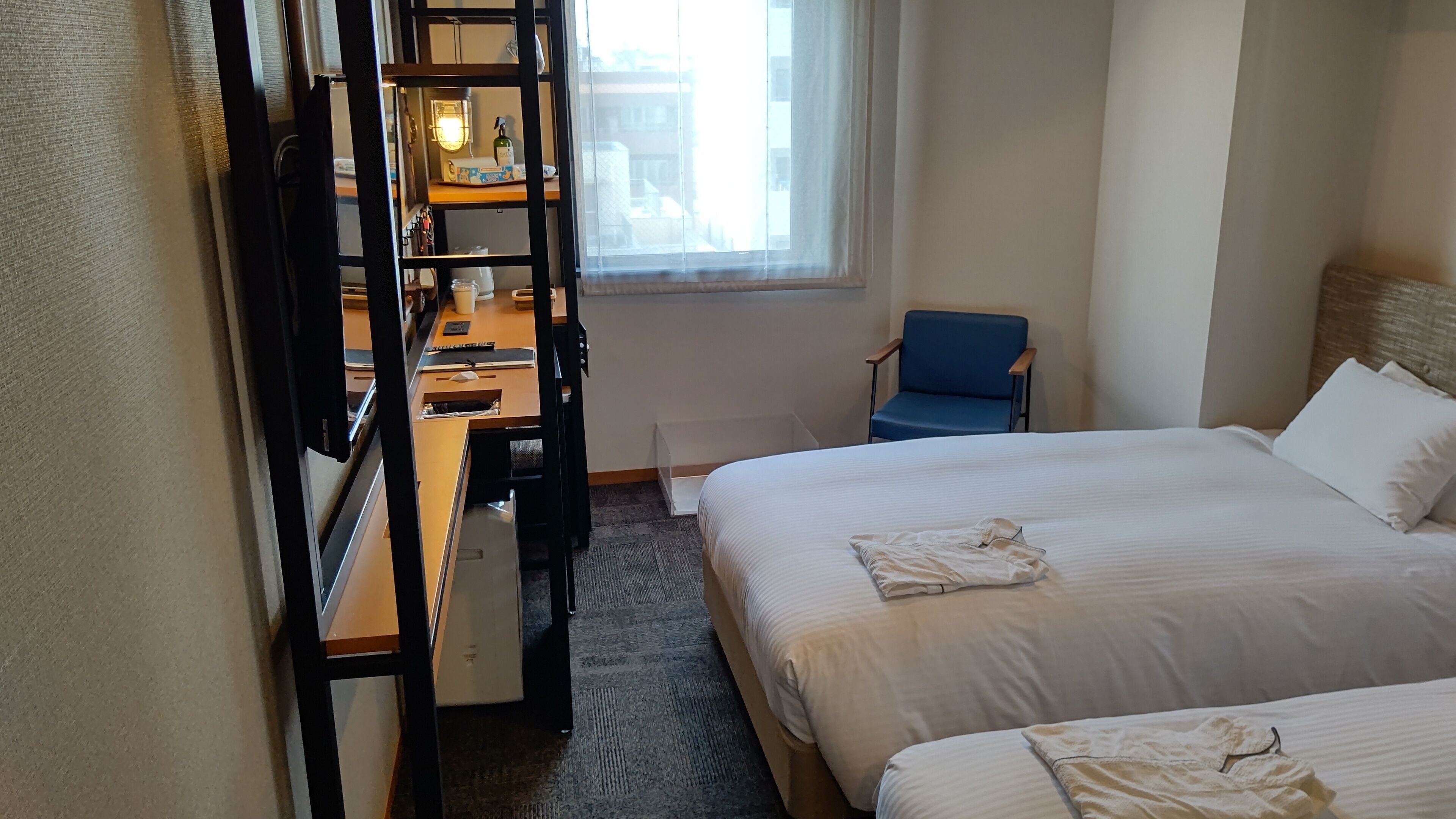 Habitación de hotel con dos camas, un escritorio con silla, una ventana con vista a edificios y una silla azul.