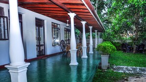 A covered outdoor area with white columns, a green floor, and a garden with chairs and potted plants. - Tamasha Mango House (Batigama)