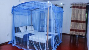 A room with a bed under a blue mosquito net, a wooden chair, a small table, a curtain, and a red floor. - Tamasha Mango House (Batigama)