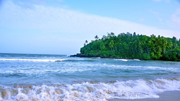 A beach with waves, a sandy shore, and a forested island in the background. - Tamasha Mango House (Batigama)