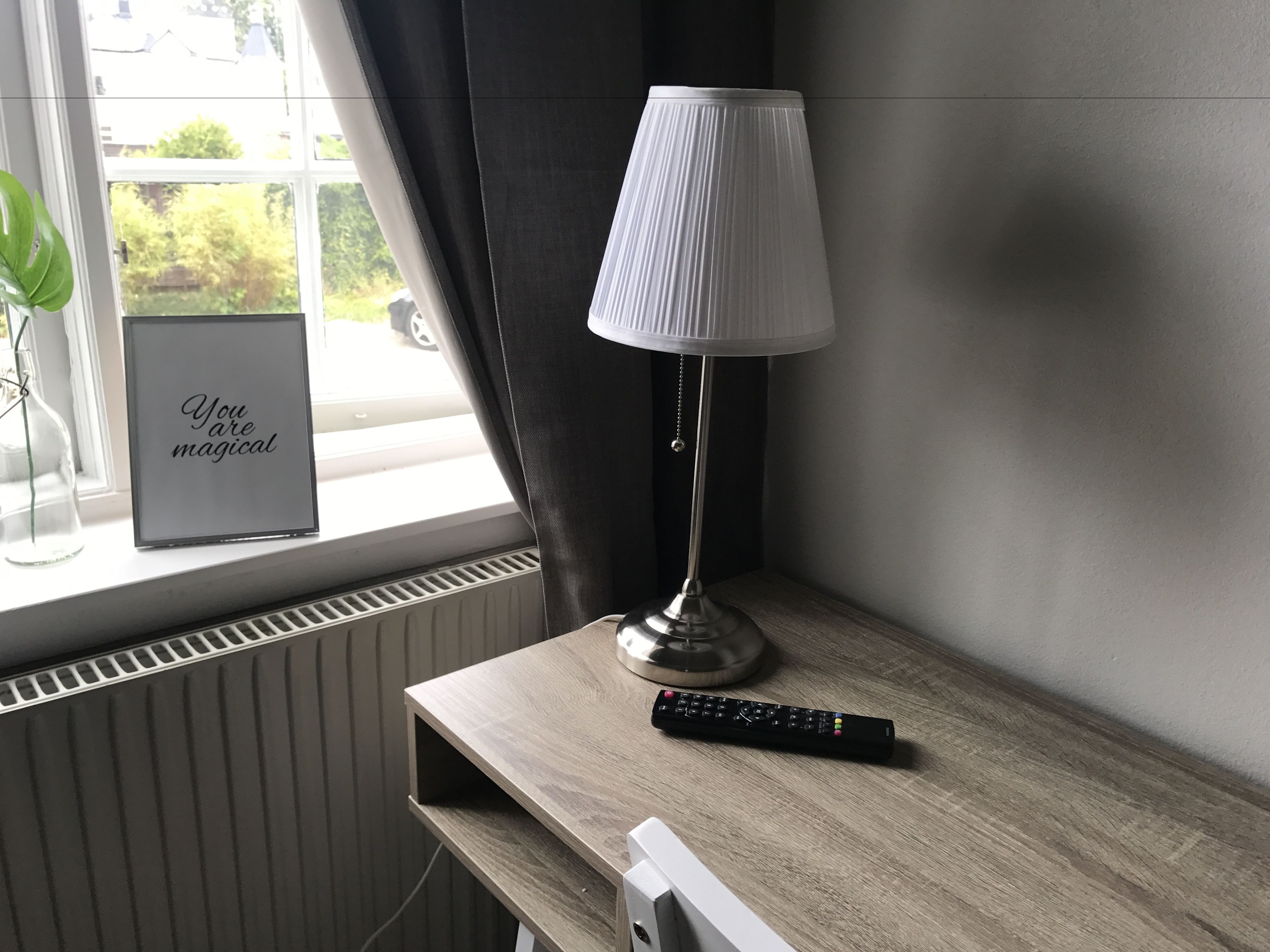 Ett nattduksbord med en lampa, en inramad tavla och en fjärrkontroll.