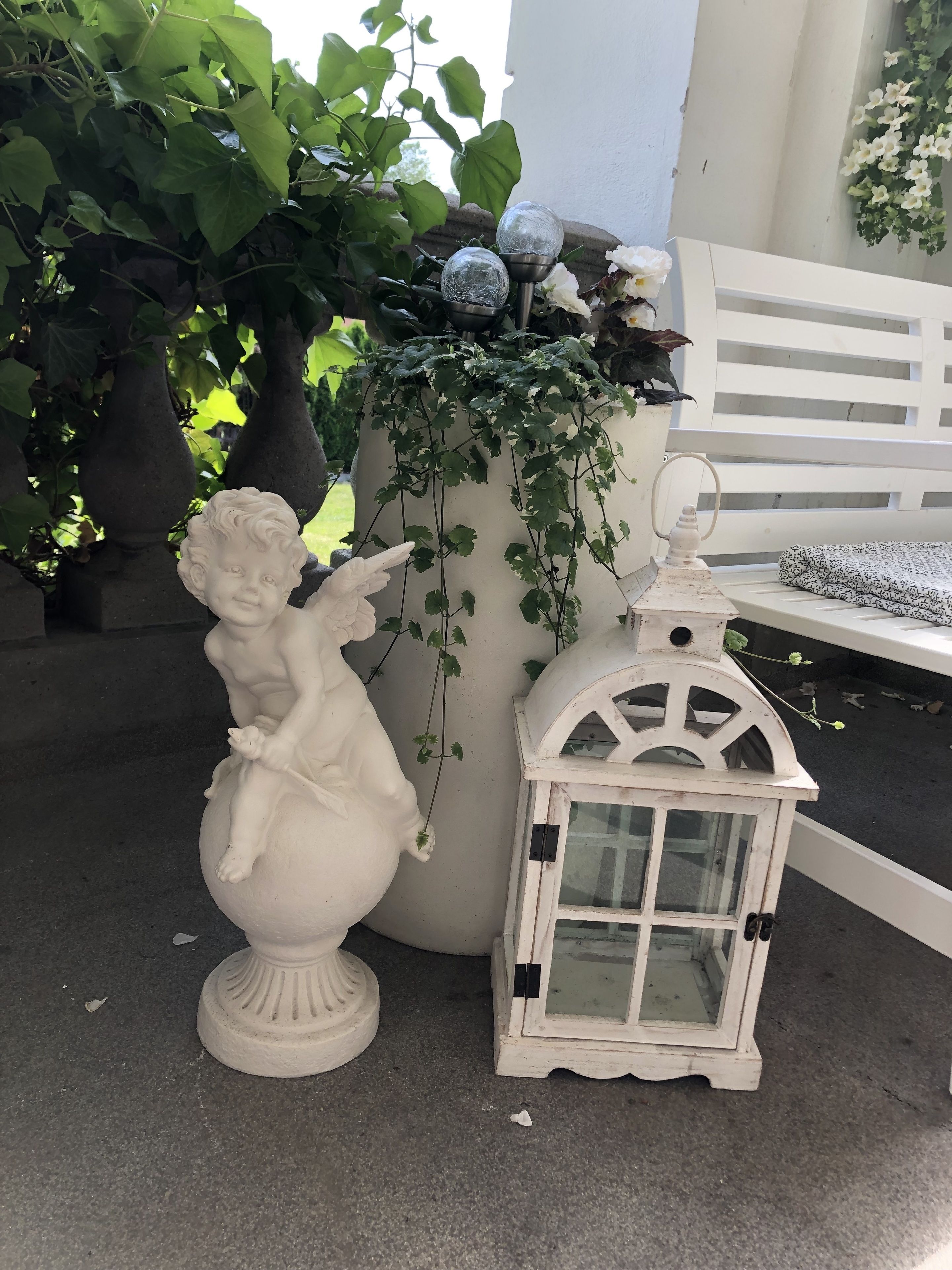 En vit utomhusstaty som föreställer en liten ängel med en katt, en vit lykta och en stor blomkruka med grönt bladverk.