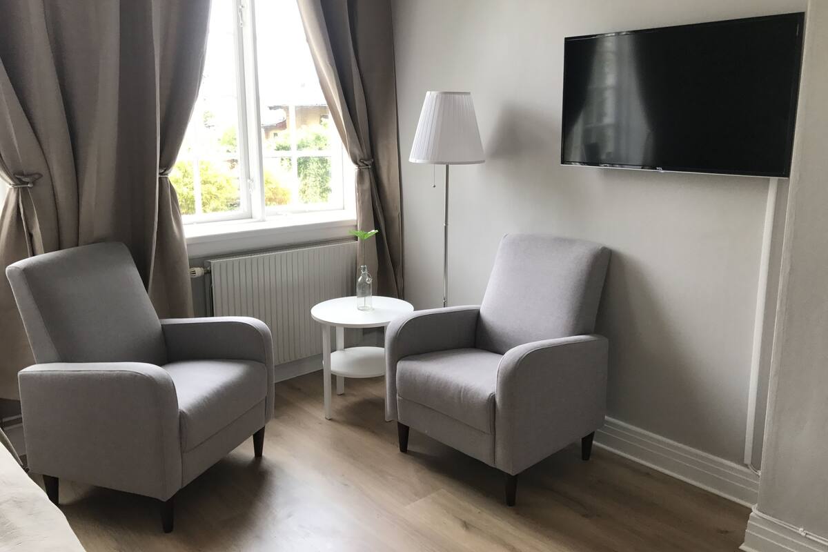 Un salon moderne avec deux fauteuils gris, une petite table blanche, un lampadaire et un téléviseur à écran plat fixé au mur.