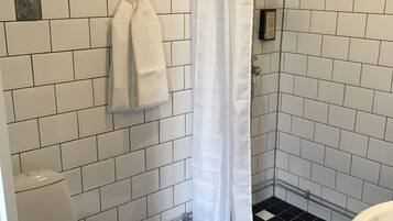 Un baño con paredes de azulejos blancos, piso a cuadros blanco y negro, inodoro blanco, ducha con cortina blanca y lavamanos blanco.