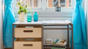 Een kamer met een raam, gordijnen, een bureau met lades, een plant en een klein tafeltje met boeken en flessen.