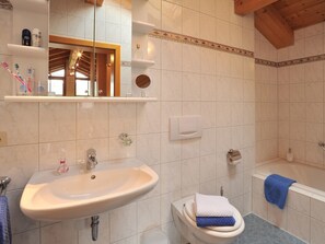 Ein Badezimmer mit Waschbecken, Toilette und Badewanne. Es gibt Regale mit Pflegeprodukten und einen Spiegel über dem Waschbecken.