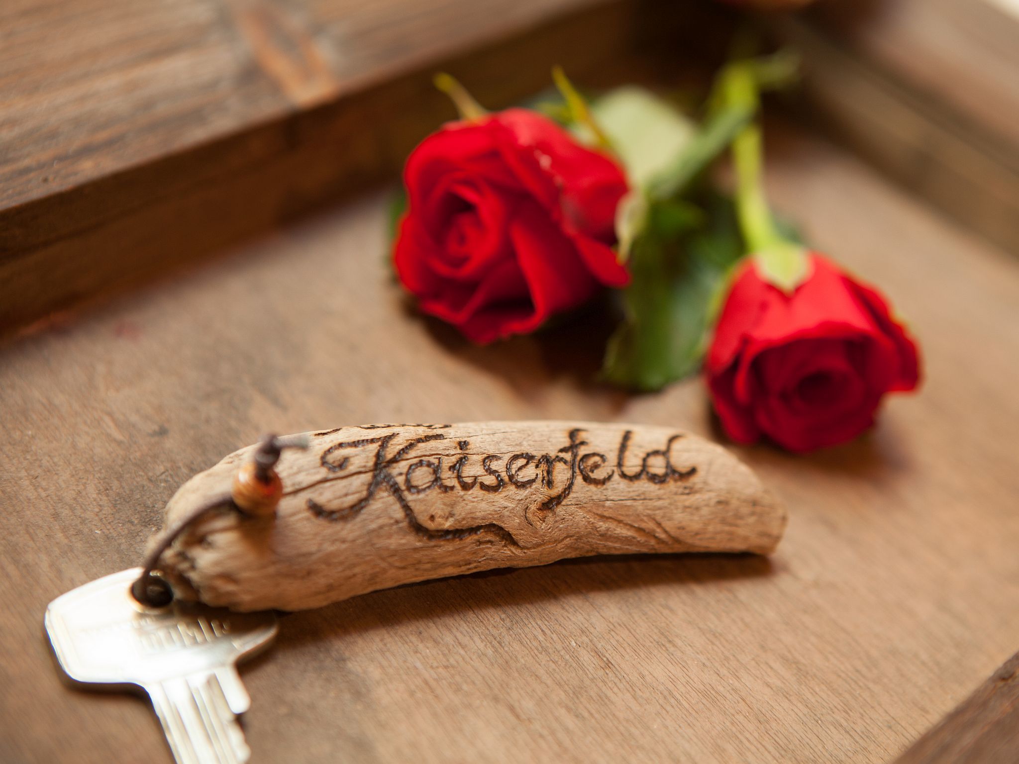 Ein Holzschlüsselanhänger mit dem Namen "Kaiserteld", auf einer Holzoberfläche liegend, mit zwei roten Rosen in der Nähe.