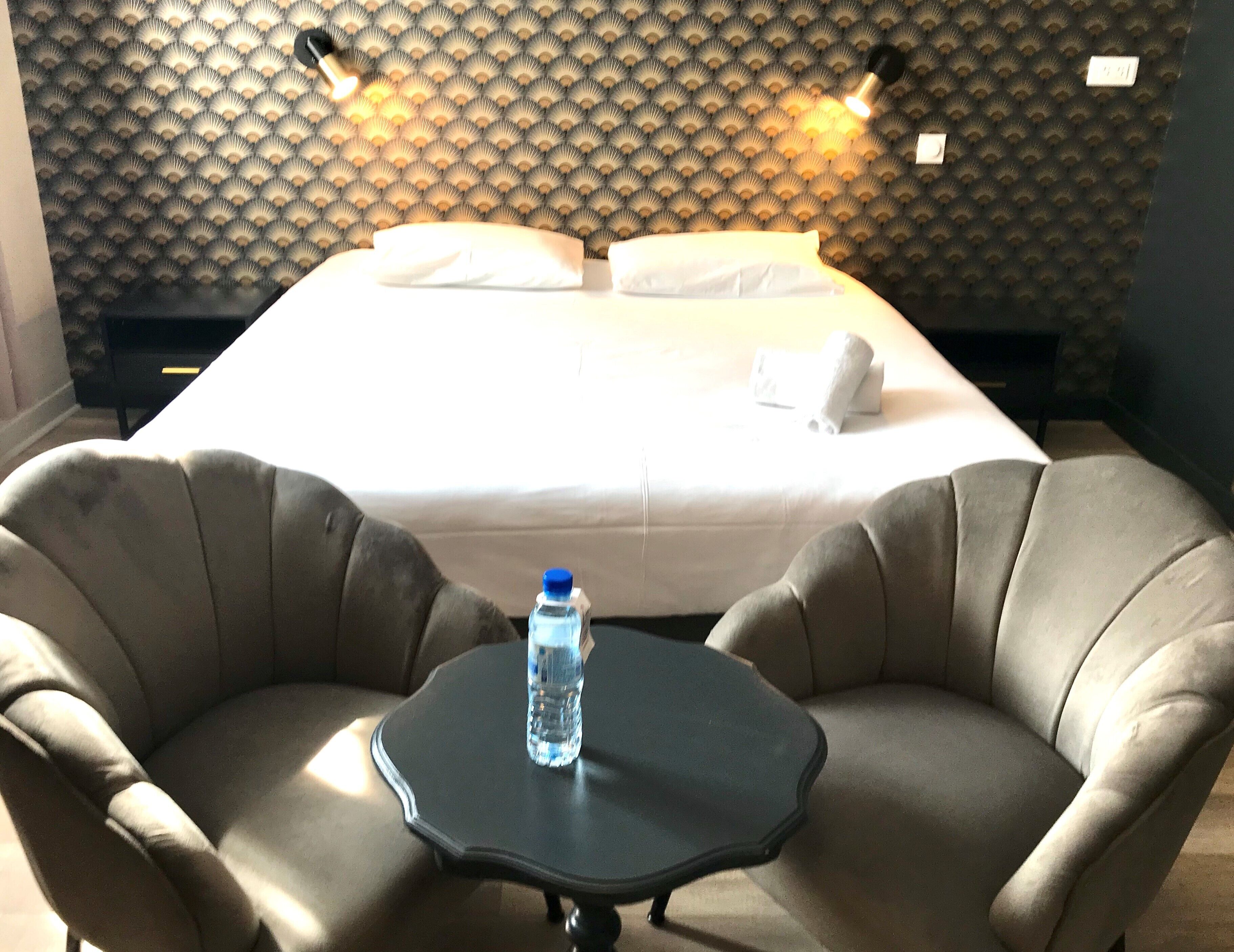 A hotel room with a bed, two armchairs, a small table, and a water bottle.