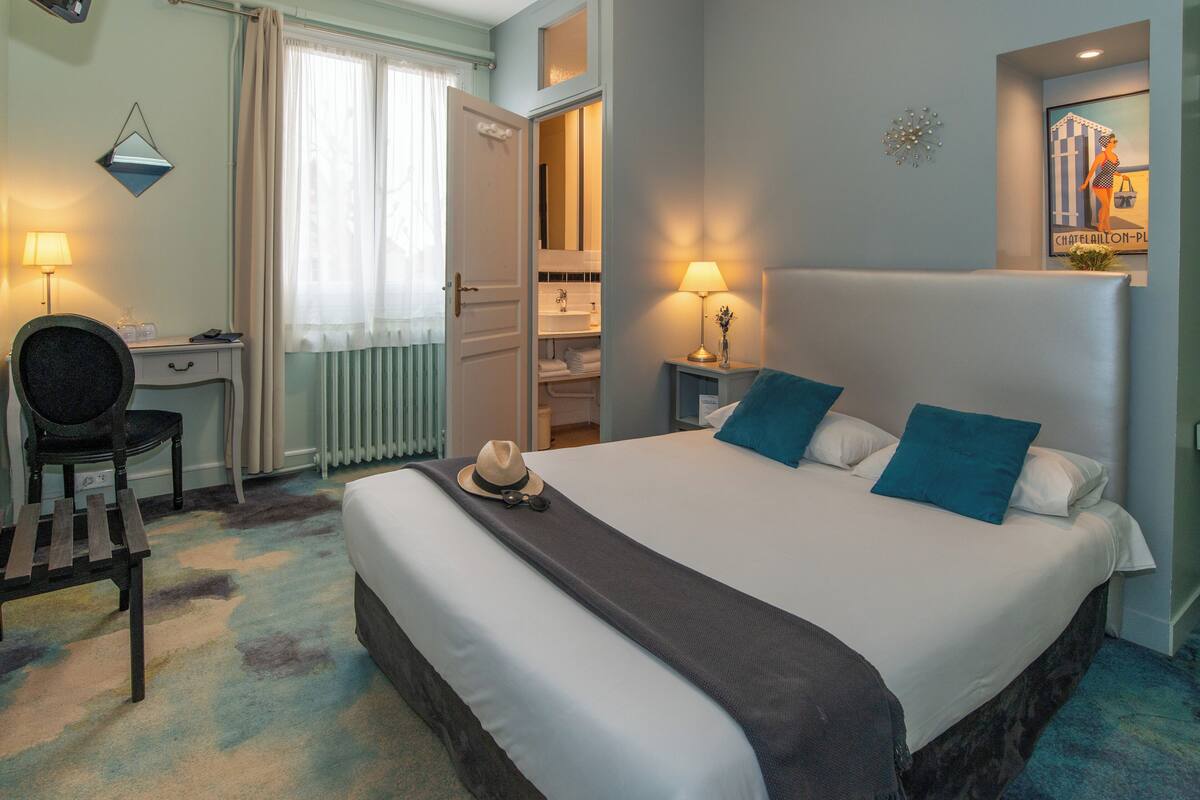 Habitación de hotel con una cama grande, un escritorio, una silla y un baño visible a través de una puerta abierta.