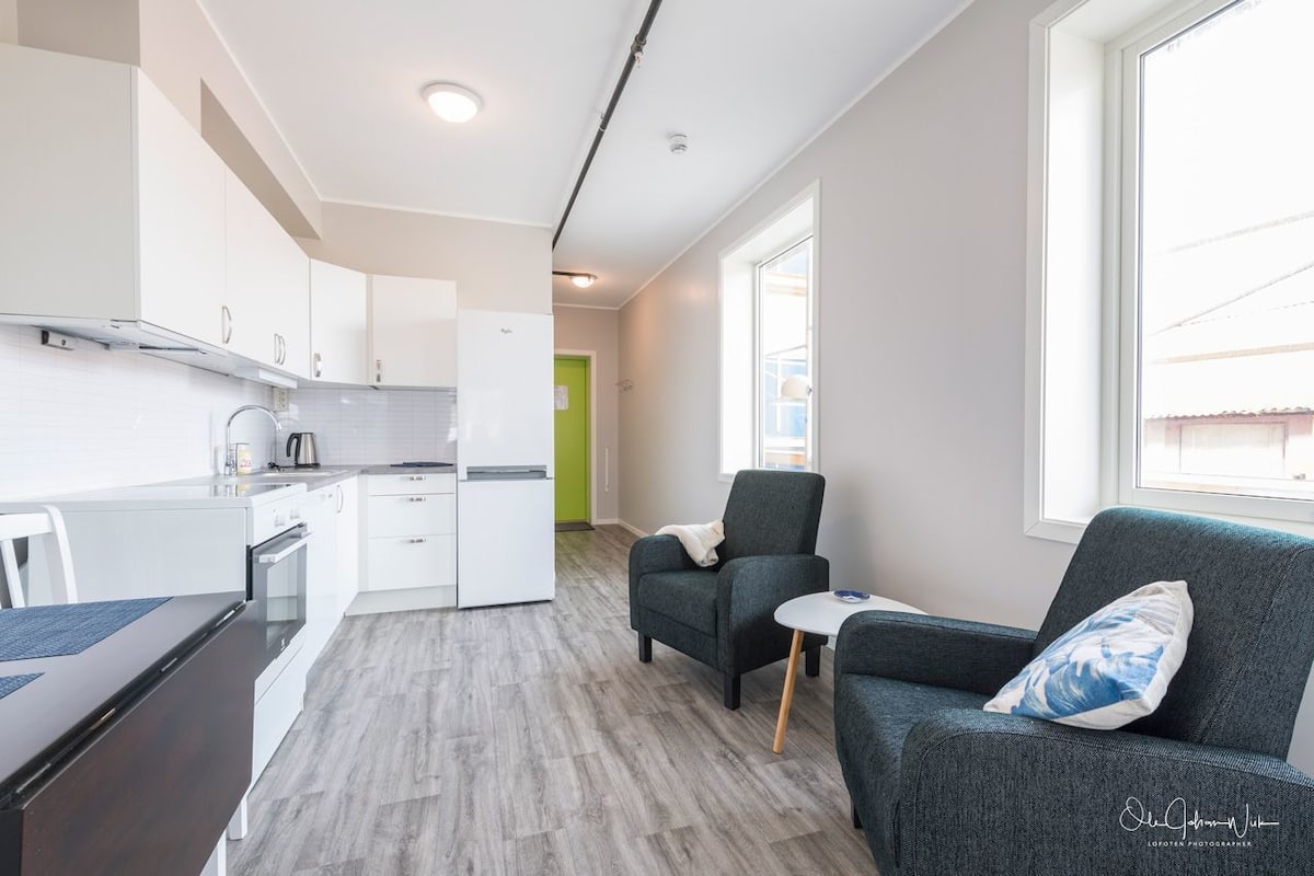Een moderne keuken met witte kasten, een inbouwoven en een groene deur naar een andere kamer. Er staat een grijze bank met een kussen en een klein wit tafeltje ernaast.