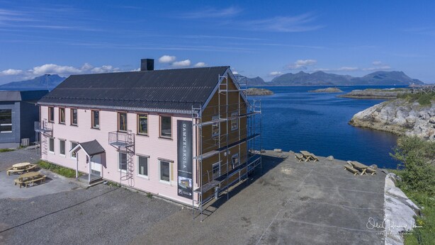 Een roze gebouw met een zwart dak, een houten terras, en uitzicht op een groot wateroppervlak en bergen op de achtergrond.