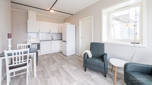 Een moderne keuken met witte kasten en een koelkast, een eethoek met een tafel en stoelen, en een grijze bank bij een raam.