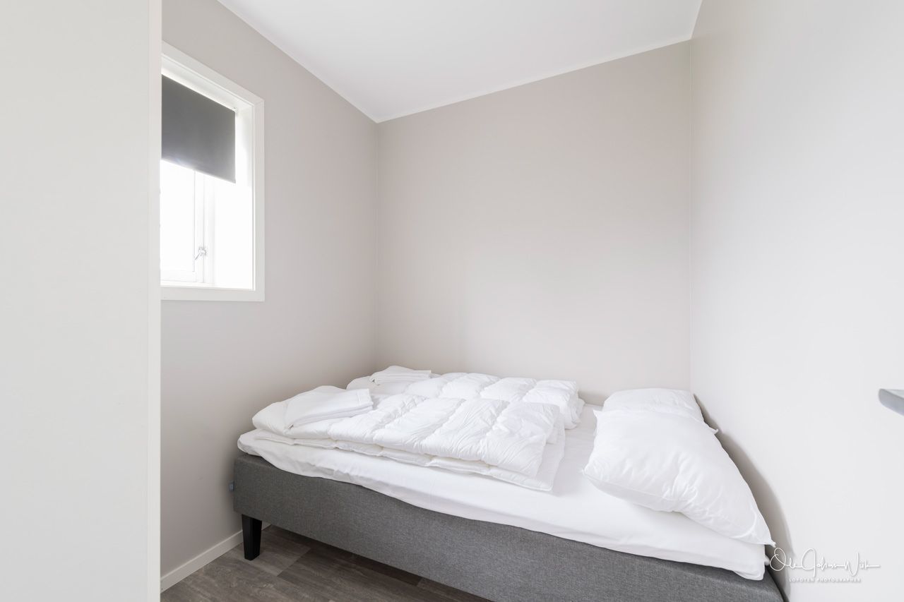 A neatly made bed in a corner room with a window, a gray bench, and a plain wall.