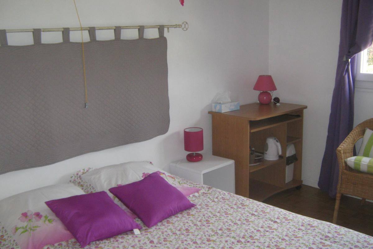Een slaapkamer met een bed, kussens, een nachtkastje, een lamp, een plank met een waterkoker en een stoel.