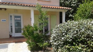 A building with a tiled roof, a door with a sign, and a garden with various plants. - Maison Venitia Chambres d'hôtes (Aussonne)