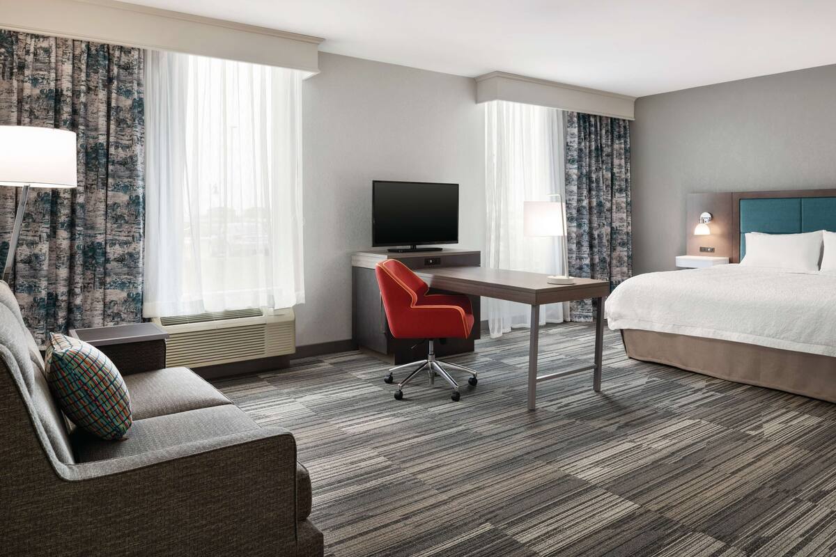 A hotel room with a bed, a desk with a TV, a red chair, and a sofa.