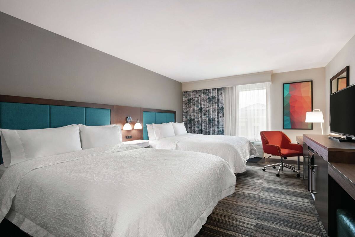 A hotel room with two beds, a desk, a chair, a TV, and a window with curtains.