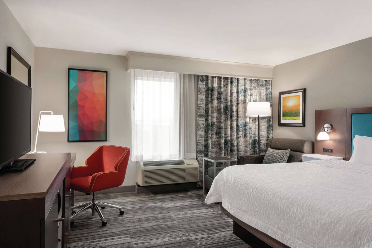 A hotel room with a bed, a desk with a chair, a TV, a window with curtains, and a wall-mounted lamp.