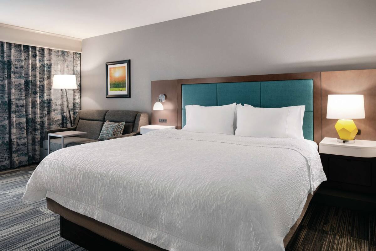 A hotel room with a large bed, a nightstand with a lamp, a seating area with a sofa and armchair, and a window with curtains.