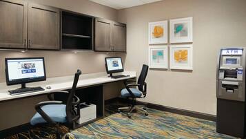 A hotel room with two desks, computers, chairs, an ATM, and framed artwork on the wall.