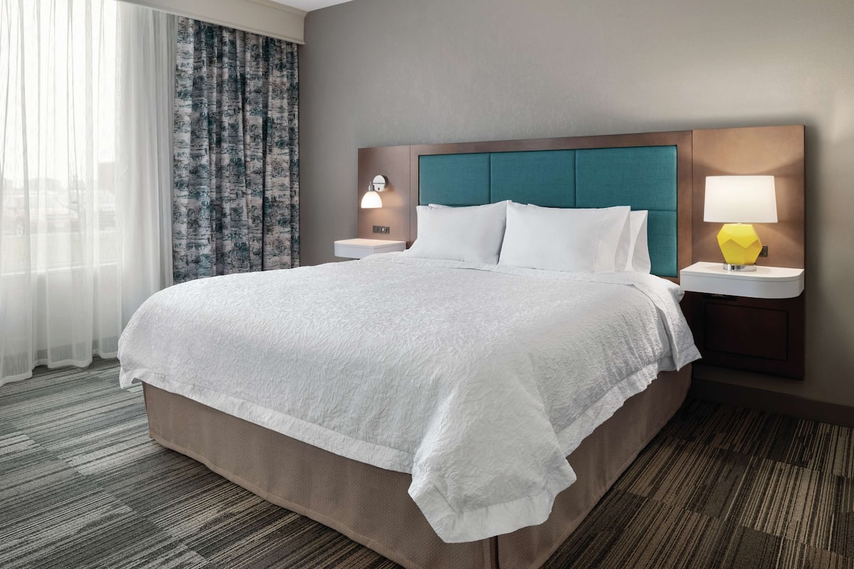 A hotel room with a large bed, two bedside tables with lamps, a window with sheer curtains, and a patterned wall.