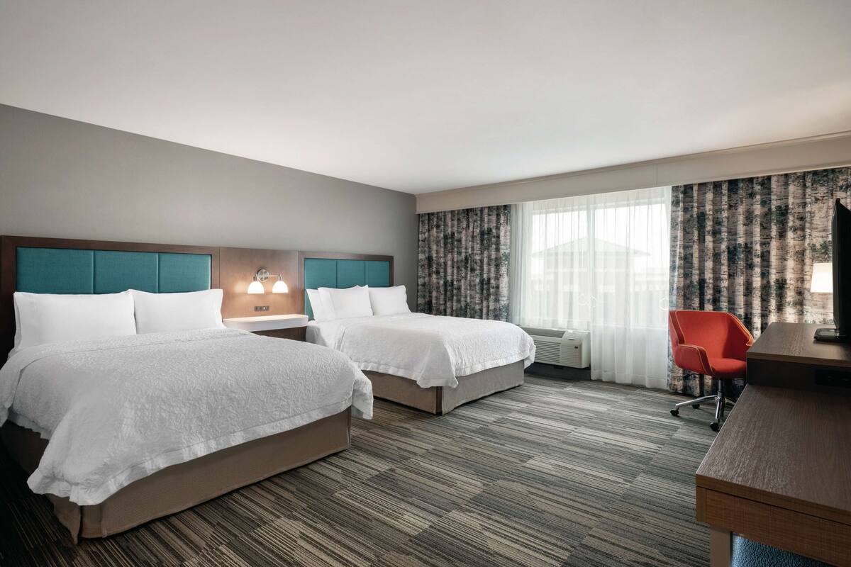 A hotel room with two beds, a desk, a chair, a lamp, and a window with curtains.