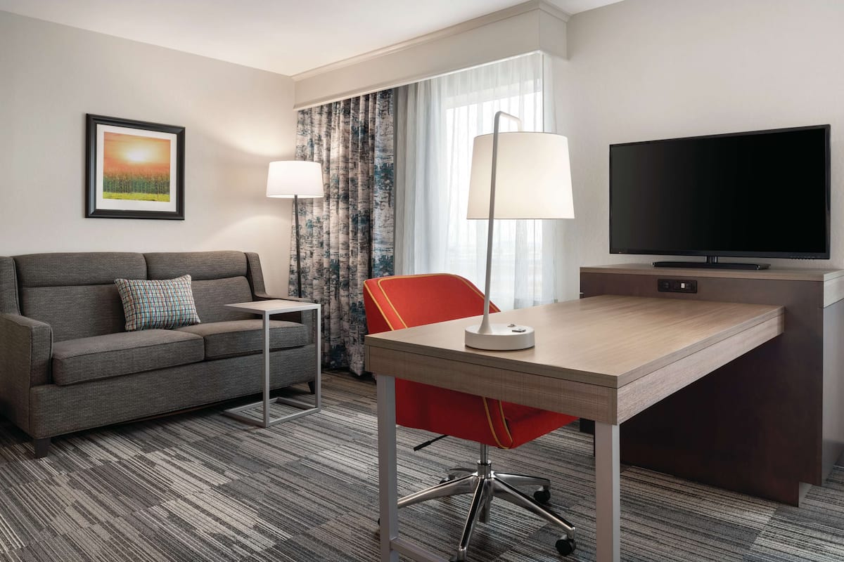 A hotel room with a grey sofa, a red chair, a wooden desk, a flat-screen TV, a lamp, and a framed picture on the wall.