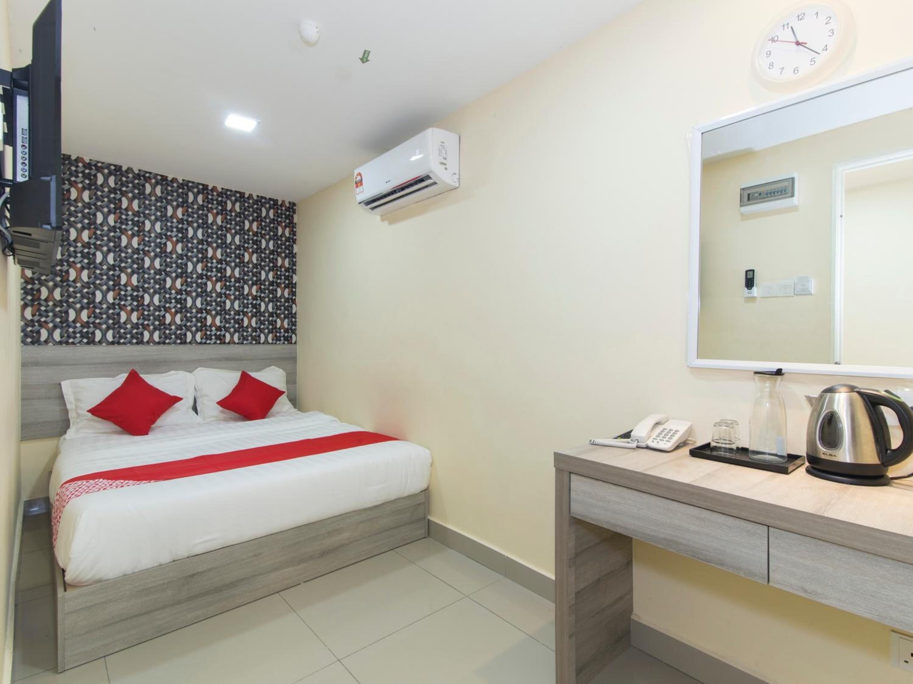 A hotel room with a bed, a desk with a phone and a kettle, a wall clock, and an air conditioning unit.