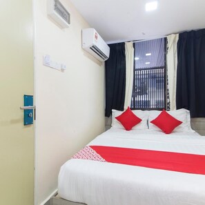 Habitación de hotel con cama, almohadas rojas, cubrecama blanco, pared azul, unidad de aire acondicionado y ventana con cortinas.