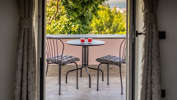 A balcony with a round table and two cushioned chairs, two glasses of red drink, and a view of green trees and buildings. - Lemon Factor Sorrento (Sorrento)