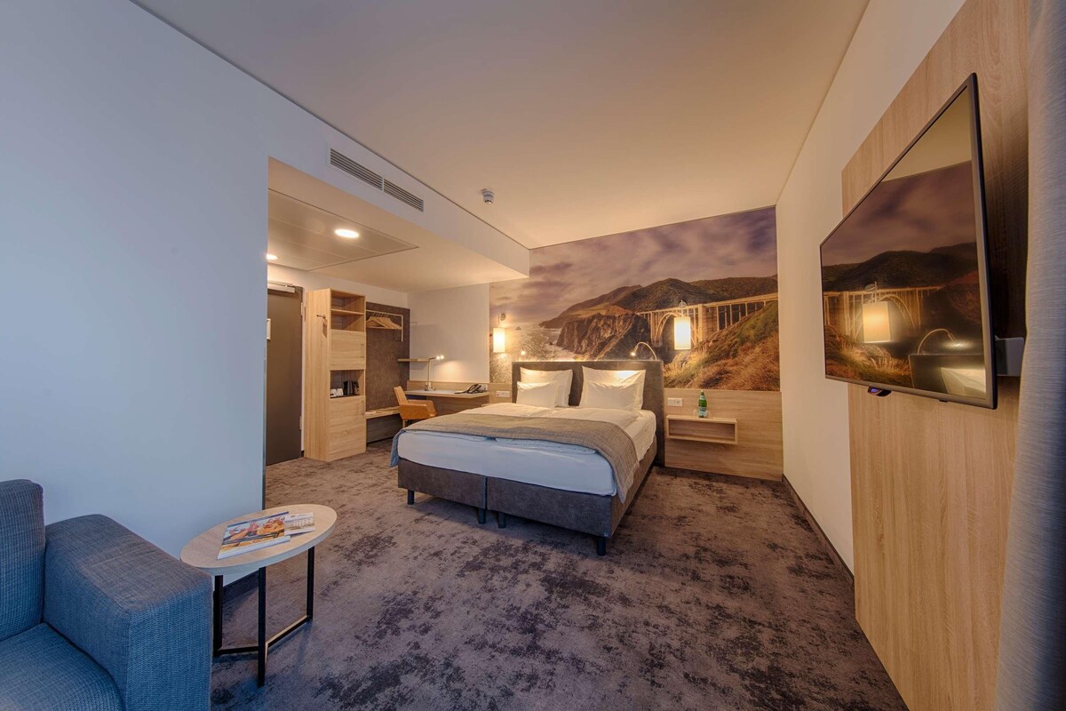 Una habitación de hotel moderna con una cama, una mesita, una silla, un escritorio y un televisor.