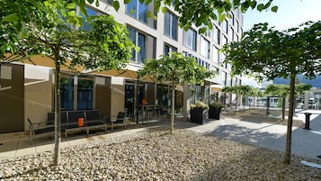 Un área de estar al aire libre con mesas y sillas, rodeada de árboles y un edificio con amplios ventanales.