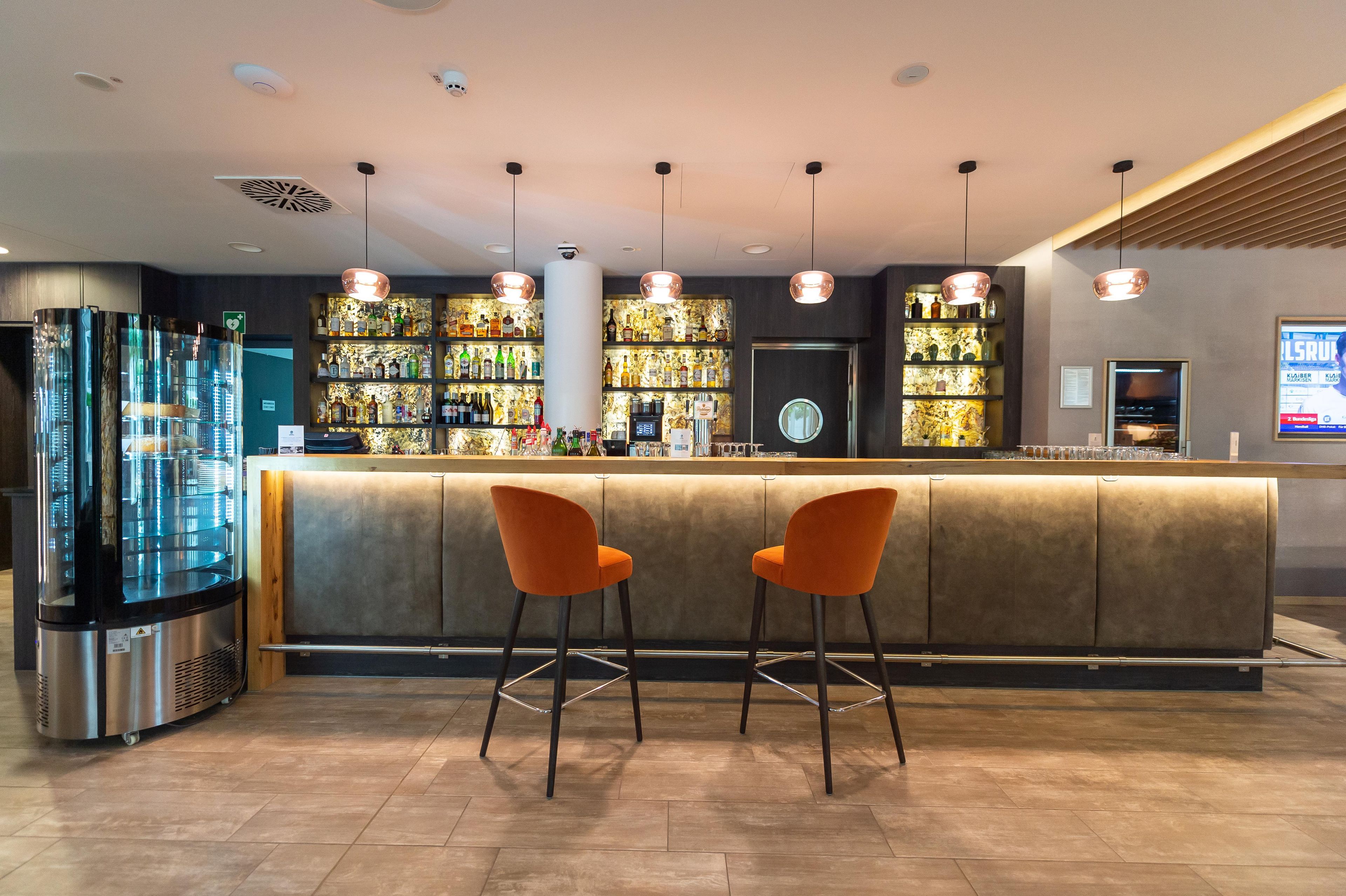 Eine Bar mit einer Theke, zwei orangefarbenen Barhocker und einem großen Kühlschrank.