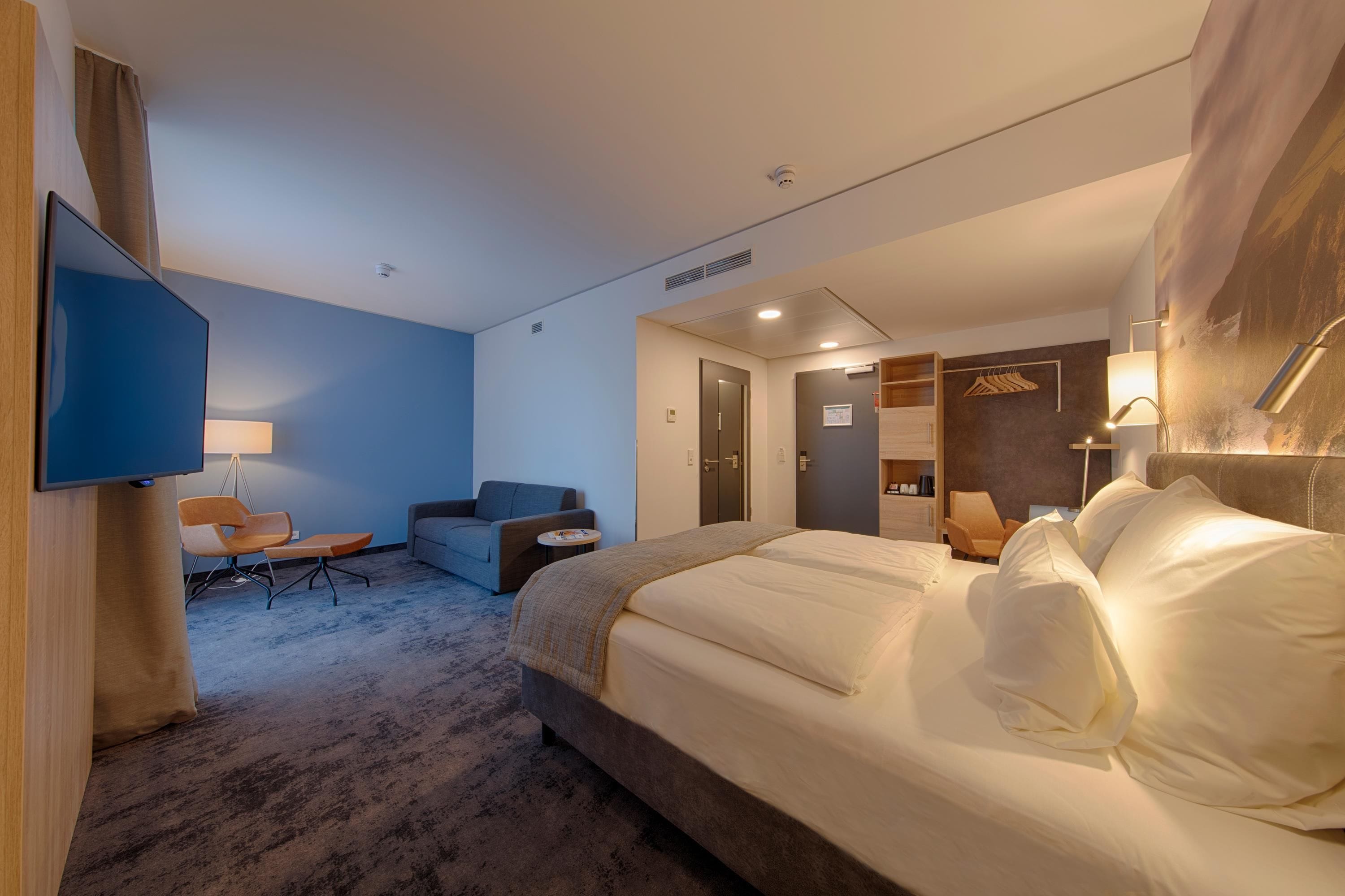 A hotel room with a large bed, a desk, a chair, a sofa, a television, and a wall-mounted light fixture.
