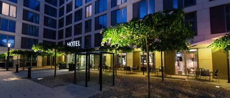 A hotel exterior with a modern design, featuring a glass facade, outdoor seating area, and a well-lit entrance.