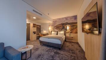 A modern hotel room with a large bed, a desk, a chair, and a small table.
