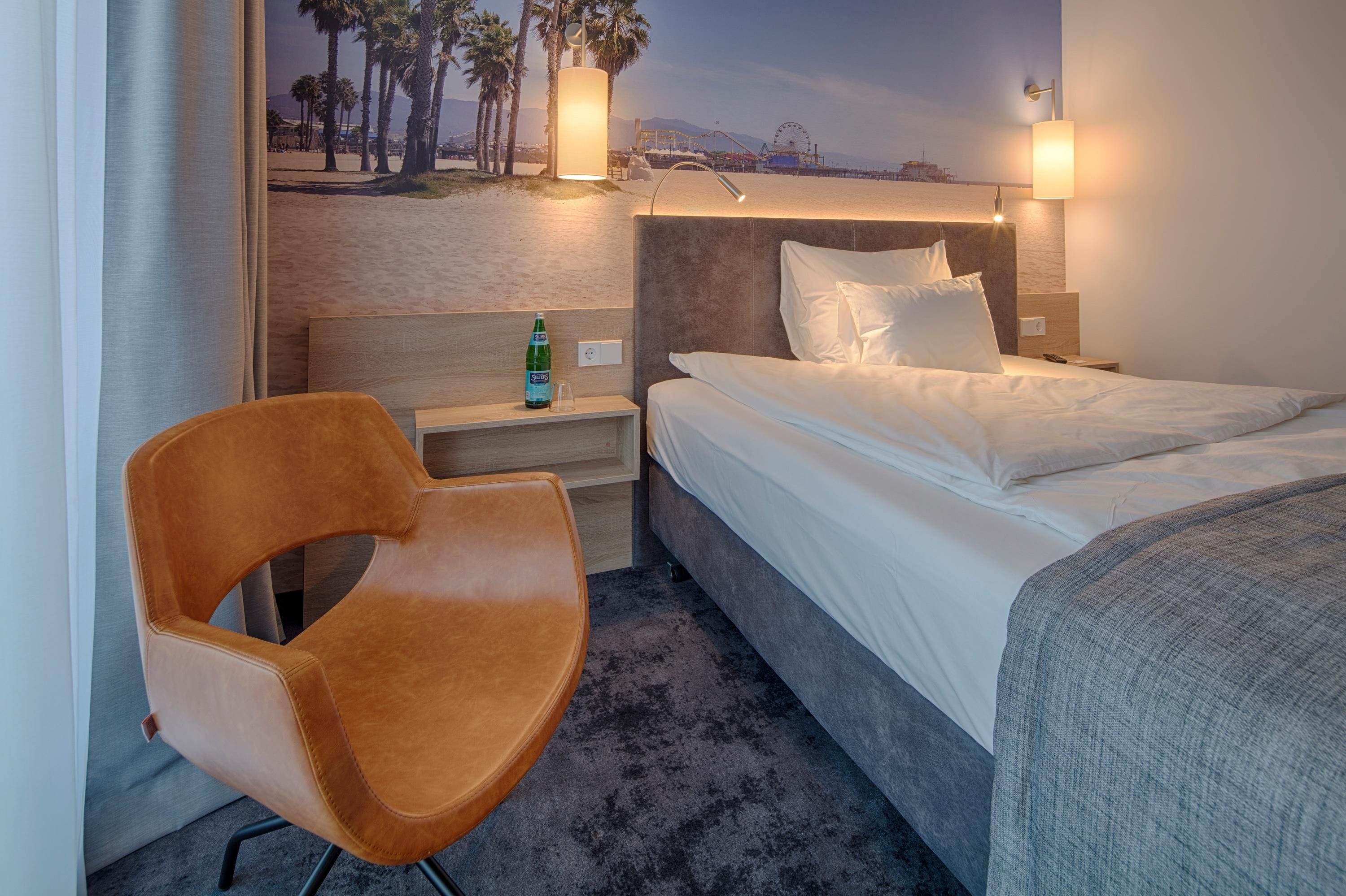 A hotel room with a bed, a chair, a nightstand, and a wall-mounted lamp.